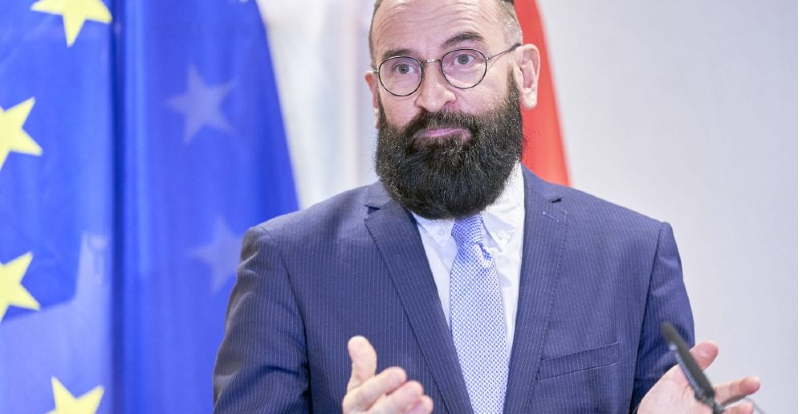 Szájer József hosszú, barna szakállal, kör alakú szemüvegben, kék zakóban, fehér ingben és világoskék nyakkendőben szónokol magyar és EU-s zászlók előtt.