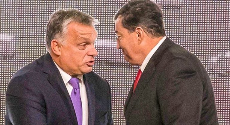 Orbán Viktor és Mészáros Lőrinc kezet fog egymással. Orbánon fekete zakó, fehér ing és lila nyakkendő van, míg Mészároson fekete zakó, fehér ing és piros nyakkendő.