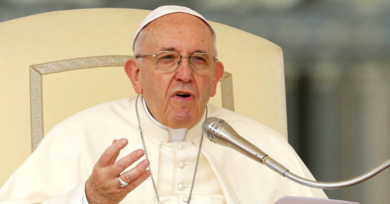 Ferenc pápa fehér ruhában ül és egy mikrofon előtt beszél a hívekhez