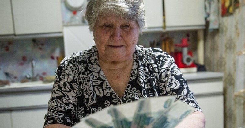 Egy idős nő sok pénzt tart a kezében a konyhájában. A nő molett, ősz hajú; elegáns fekete-fehér ruhát visel.