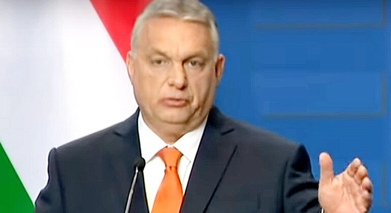 Orbán Viktor fekete zakóban, fehér ingben, narancssárga nyakkendőben, ősz hajjal és gondterhelt tekintettel beszél a magyar zászló előtt.