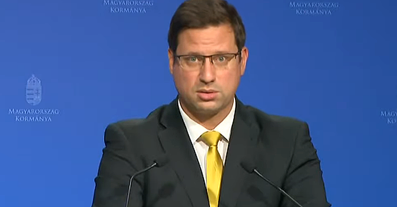 Gulyás Gergely miniszter kék háttér előtt sötét zakóban szemüvegben és sárga nyakkendőben beszél