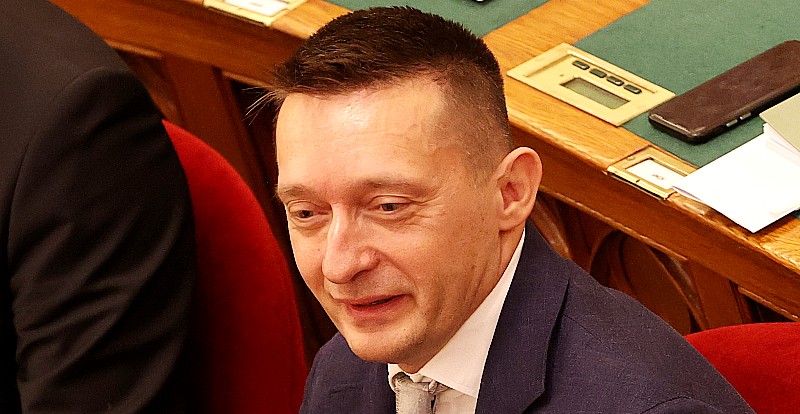 Rogán Antal vörös bársonyszékben, kék zakóban, fehér ingben és szürke nyakkendőben ül a parlamentben, és furcsa fejet vág.