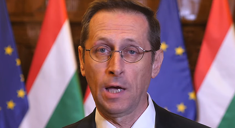 Varga mihály pénzügyminiszter szemüvegben zászlók előtt tátott szájjal áll és beszél
