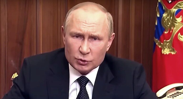 Vlagyimir Putyin fekete zakóban, fehér ingben, fekete nyakkendőben, idegesen nézve beszél.