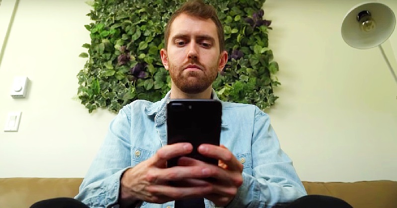 Egy telefonját néző férfi kék ingben egy fehér fal előtt ül.