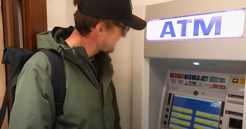 ATM, ügyfél fekete sapkában szemüvegben, zöld kabátban, hátizsákkal MBH