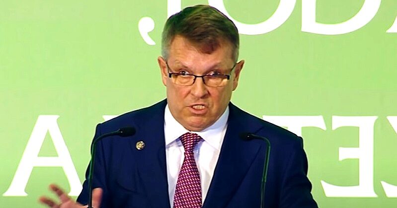 Matolcsy György zöld háttér előtt, sötétkék öltönyben, lila nyakkendőben, beszédet mond