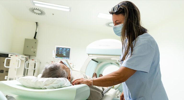 A képen egy MR-készülék előtt fekvő páciens látható a Semmelweis Egyetem egyik kórházában. Egy fehér köpenyes, barna hajú, maszkos asszisztens nyugtatja őt.