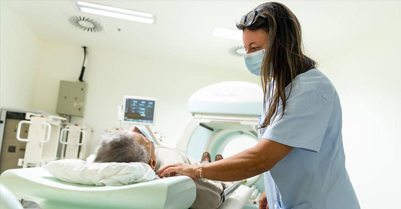 A képen egy MR-készülék előtt fekvő páciens látható a Semmelweis Egyetem egyik kórházában. Egy fehér köpenyes, barna hajú, maszkos asszisztens nyugtatja őt.