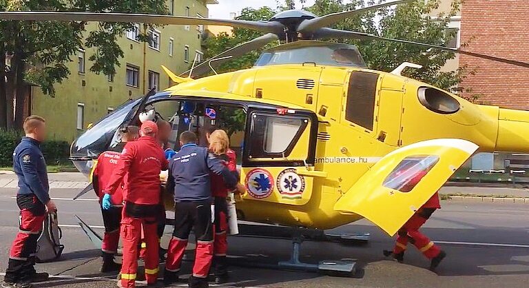Sárga színű mentőhelikopterbe szállnak be az Országos Mentőszolgálat munkatársai egy zöld színű panelház mellett.