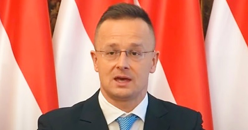 Szijjártó Péter zászló előtt áll szemüvegben fekete zakóban kék nyakkendőben és fehér ingben. Újságíróknak nyilatkozik a miniszter