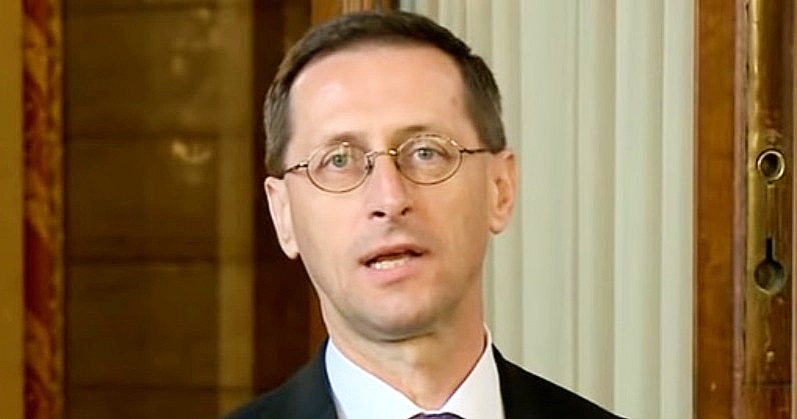 Varga Mihály fekete zakóban, fehér ingben, nyakkendőben és kör alakú szemüvegben néz a kamerába.