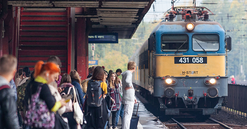 Szili típusú villanymozdony érkezik szerelvényével Kőbánya-Kispest vasútállomásra. A vonat hosszú, a várakozó tömeg nagy. A vonat kék és sárga színű.