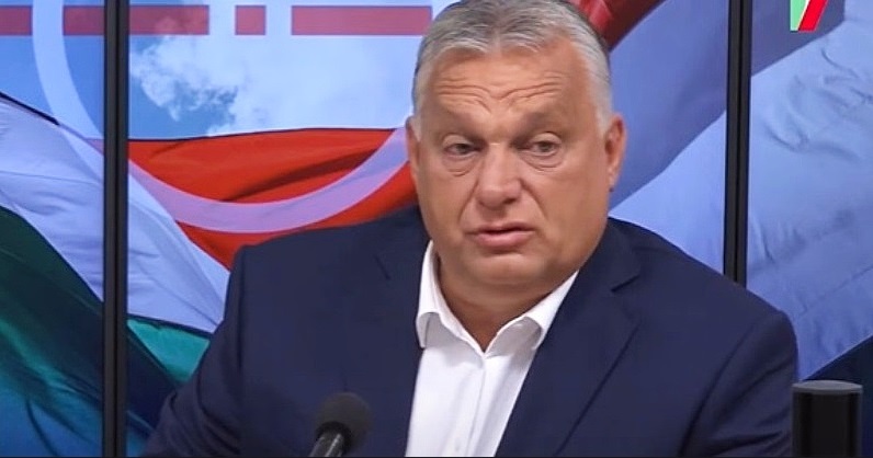 Orbán Viktor sötét öltönyben ül fehér ingben egy stúdióba és interjút ad nemzeti színű zászlók előtt és furcsa fejet vág