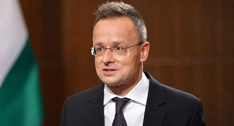 Szijjártó Péter külgazdasági és külügyminiszter a magyar zászló előtt fekete öltönyben, fehér ingben