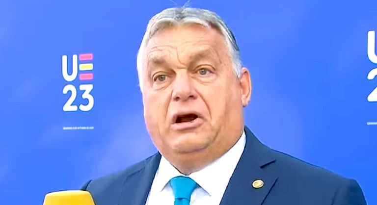 Orbán Viktor sötétkék zakóban, fehér ingben, világoskék nyakkendőben, kitűzőben, ősz hajjal, ráncos homlokkal furcsán néz.