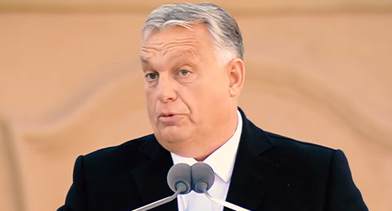 Orbán Viktor fekete zakóban, fehér ingben beszél a mikrofonokba. Haja ősz, homloka ráncos.