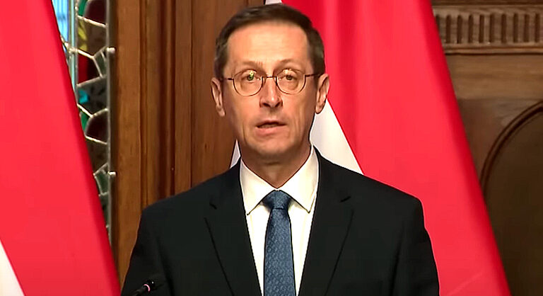 Varga Mihály szemüvegben zászló előtt sötét zakóban fehér ingben kék nyakkendővel beszél