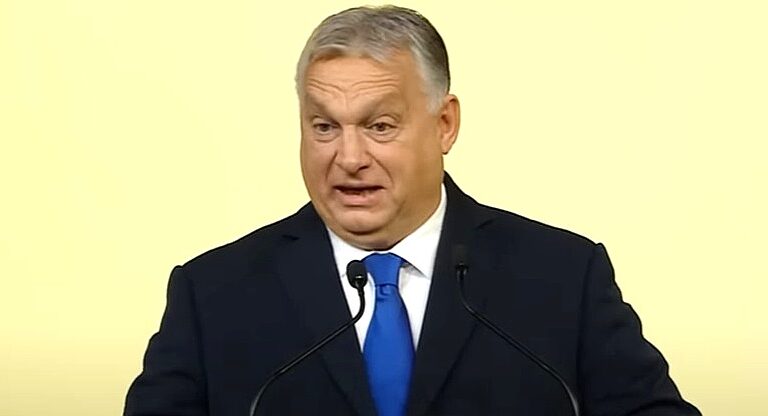 Orbán Viktor sárga háttér előtt, fekete öltönyben, fehér ingben, kék nyakkendőt visel