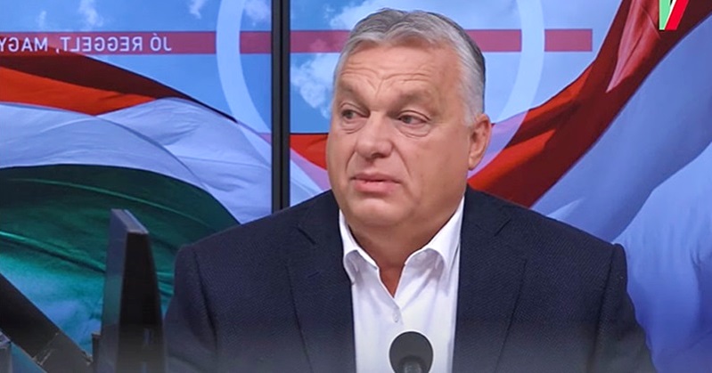 Orbán Viktor beszél egy stúdióban zászlós háttér előtt sötét zakóban és fehér ingben
