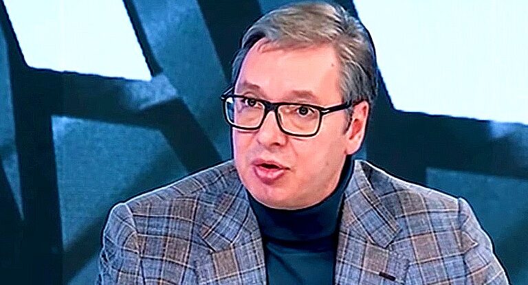 Aleksandar Vučić kockás zakóban, feketekeretes szemüvegben, fekete, garbós pulcsiban beszél a kamerába egy stúdióban.