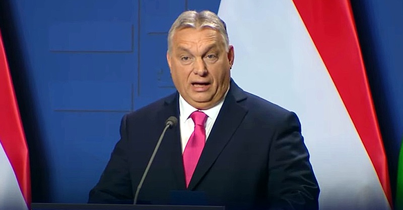 Orbán Viktor kék háttér előtt fekete öltönyben, fehér ingben, rózsaszín nyakkendőben nyilatkozik.