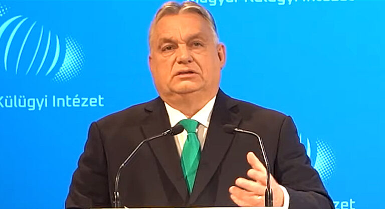 Orbán Viktor fekete zakóban, fehér ingben, zöld nyakkendőben a kamerába nézve mutogat.