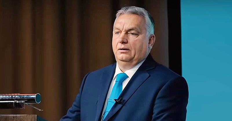 Orbán Viktor sötétkék öltönyben, fehér ingben, kék nyakkendőben a pódiumon, mikrofonok előtt