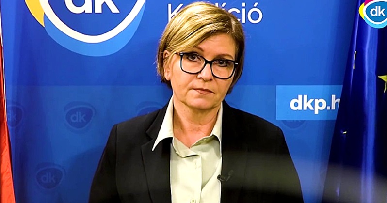 Kálmán Olga sajtótájékoztatót tart kék háttér előtt fehér ingben fekete kabátban