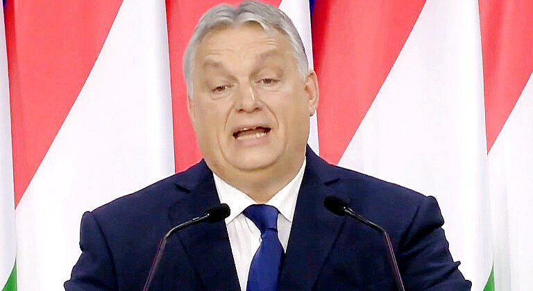 Orbán Viktor magyar zászlók előtt beszél kék színű zakóban, fehér ingben és kék nyakkendőben, ősz hajjal, miközben furcsán grimaszol. Előtte két mikrofon van.