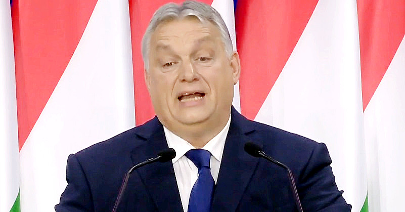 Orbán Viktor magyar zászlók előtt beszél kék színű zakóban, fehér ingben és kék nyakkendőben, ősz hajjal, miközben furcsán grimaszol. Előtte két mikrofon van.