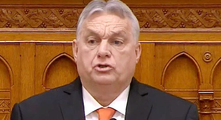 Orbán Viktor a parlamentben beszél ráncos, meggyötört fejjel, fekete zakóban, fehér ingben és narancssárga nyakkendőben.