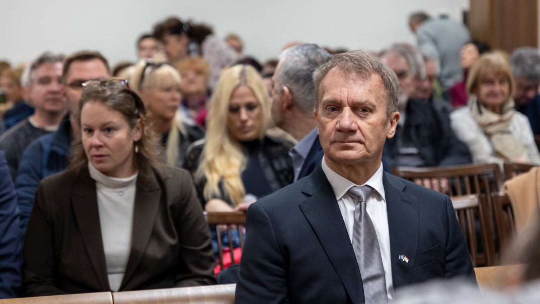 Varju László a tárgyalóteremben ül, előre néz. Rajta fekete zakó, fehér ing és szürke nyakkendő látható. Mögötte a támogatói ülnek.