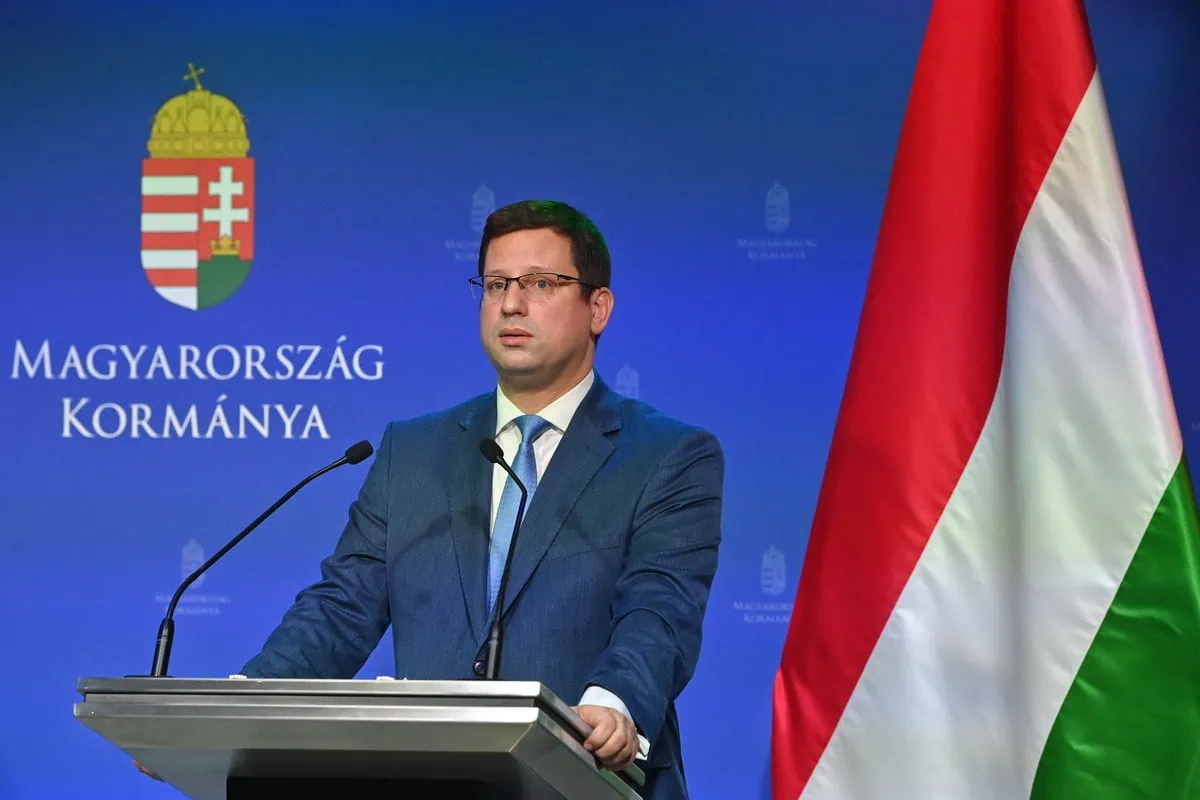 Gulyás Gergely kormányinfót tart kék háttér előtt magyar zászló mellett szemüvegben sötét zakóban