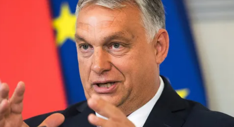 Orbán Viktor mutogat az EU zászlaja előtt ősz hajjal, fekete zakóban és fehér ingben.