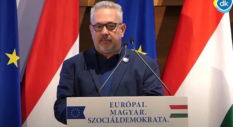 Arató Gergely, magyar zászló, EU-s zászló, mikrofon, sötétkék zakó, sötétkék ing