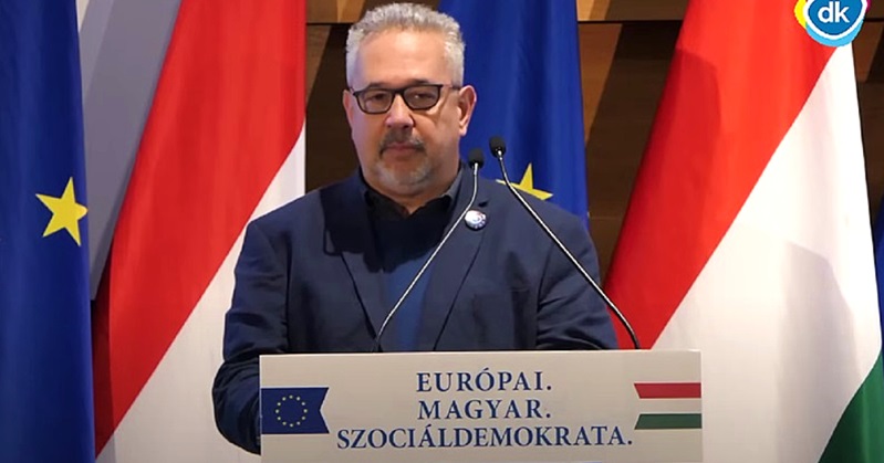 Arató Gergely, magyar zászló, EU-s zászló, mikrofon, sötétkék zakó, sötétkék ing