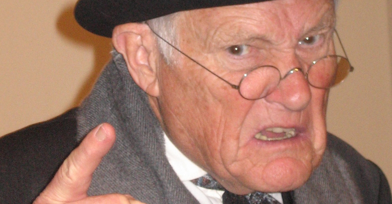 Dühös idős férfi szemüvegben szürke sálban, fehér ingben,fekete kalapban mutat