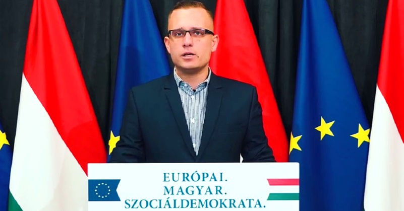 Mészáros Dávid, a Demokratikus Koalíció politikusa sötétkék öltönyben, kék ingben, a pódiumon mikrofonok előtt sajtótájékoztatót tart.