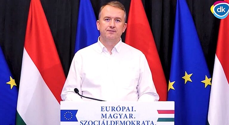 Sebián-Petrovszki László, a DK politikusa a magyar és az EU-s zászló előtt, mikrofonban, fehér inget viselve nyilatkozik.