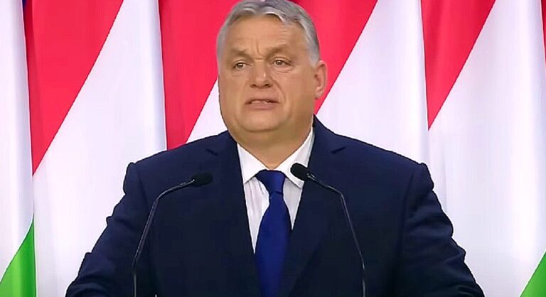 Orbán Viktor zászlók előtt áll sötétkék öltönyben, kék nyakkendőben, mikrofonok előtt