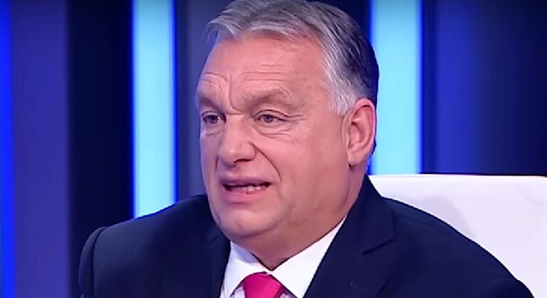 Orbán Viktor kék háttér előtt, sötétkék öltönyben, piros nyakkendőt visel, interjút ad egy stúdióban