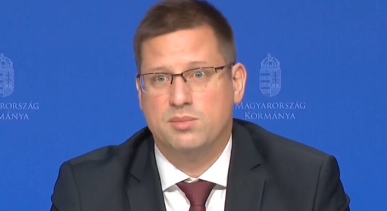 Gulyás Gergely megdöbbenten néz a kormányinfót. Rajta fekete zakó, fehér ing és vörös nyakkendő látható.