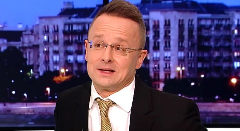 Szijjártó Péter fekete öltönyben ad interjút az egyik tévéstúdióban, nyakkendőt és szemüveget visel, fehér ingben van és éppen beszél