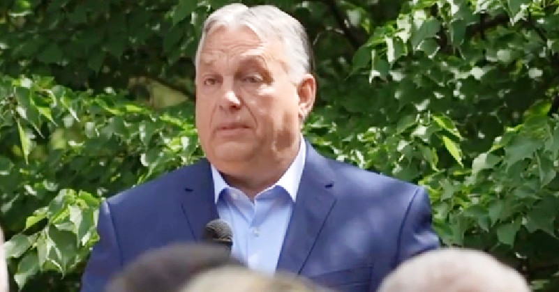 Orbán Viktor sötétkék zakóban, világoskék ingben a mikrofonba beszél néhány ember előtt a természetben.