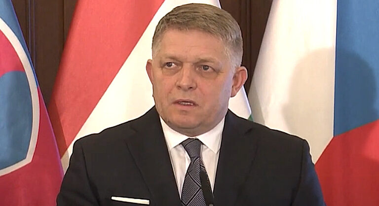 Robert Fico fekete zakóban. fehér ingben, fekete nyakkendőben szlovák zászló előtt beszél, miközben balra néz.