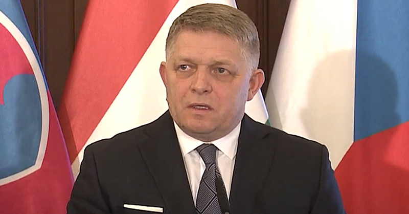 Robert Fico fekete zakóban. fehér ingben, fekete nyakkendőben szlovák zászló előtt beszél, miközben balra néz.
