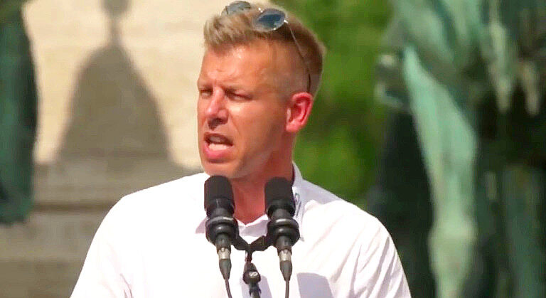 Magyar Péter fehér ingben, vörös fejjel, idegesen beszél két mikrofon előtt, miközben balra fordul.