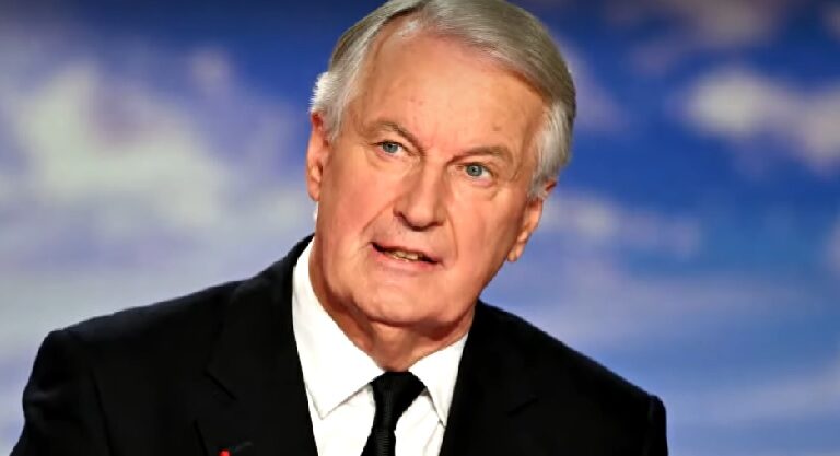 Michel Barnier fekete zakóban, fehér ingben, fekete nyakkendőben, ősz jajjal, balra nézve beszél.